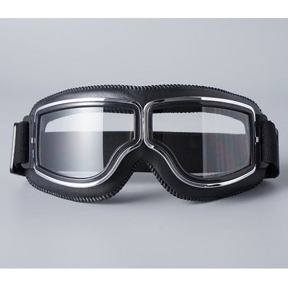 Front view of the black-padded goggles with clear lenses, chrome trim and a black elastic strap on a grey background. 16