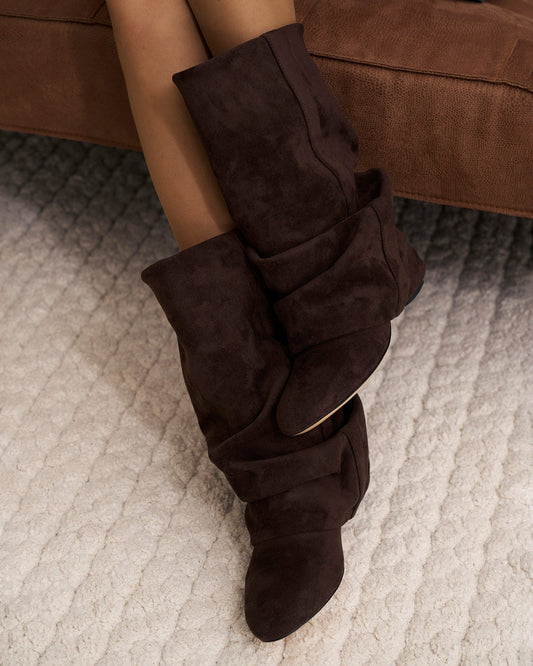 Close-up of a model's legs in sheer tights wearing the slouchy boots, seated on a brown sofa above a cream rug. 2