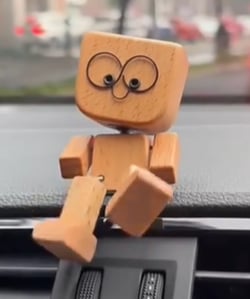 Close-up of the wooden figure with round wire glasses face, sitting on a car dashboard with one leg hanging down. 6