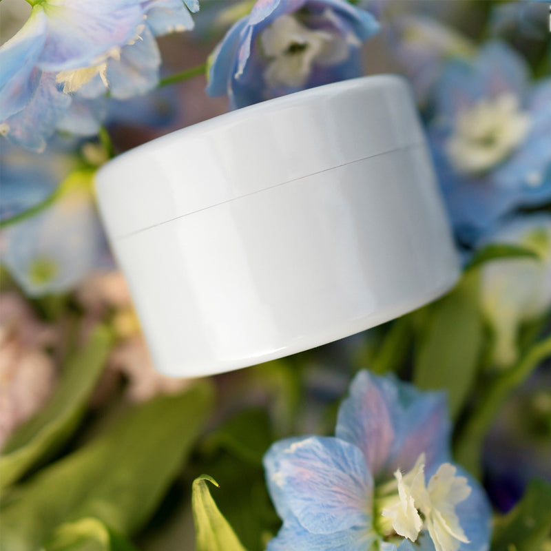 Closed white round insert lying on a bed of blue delphinium flowers and green leaves. 9