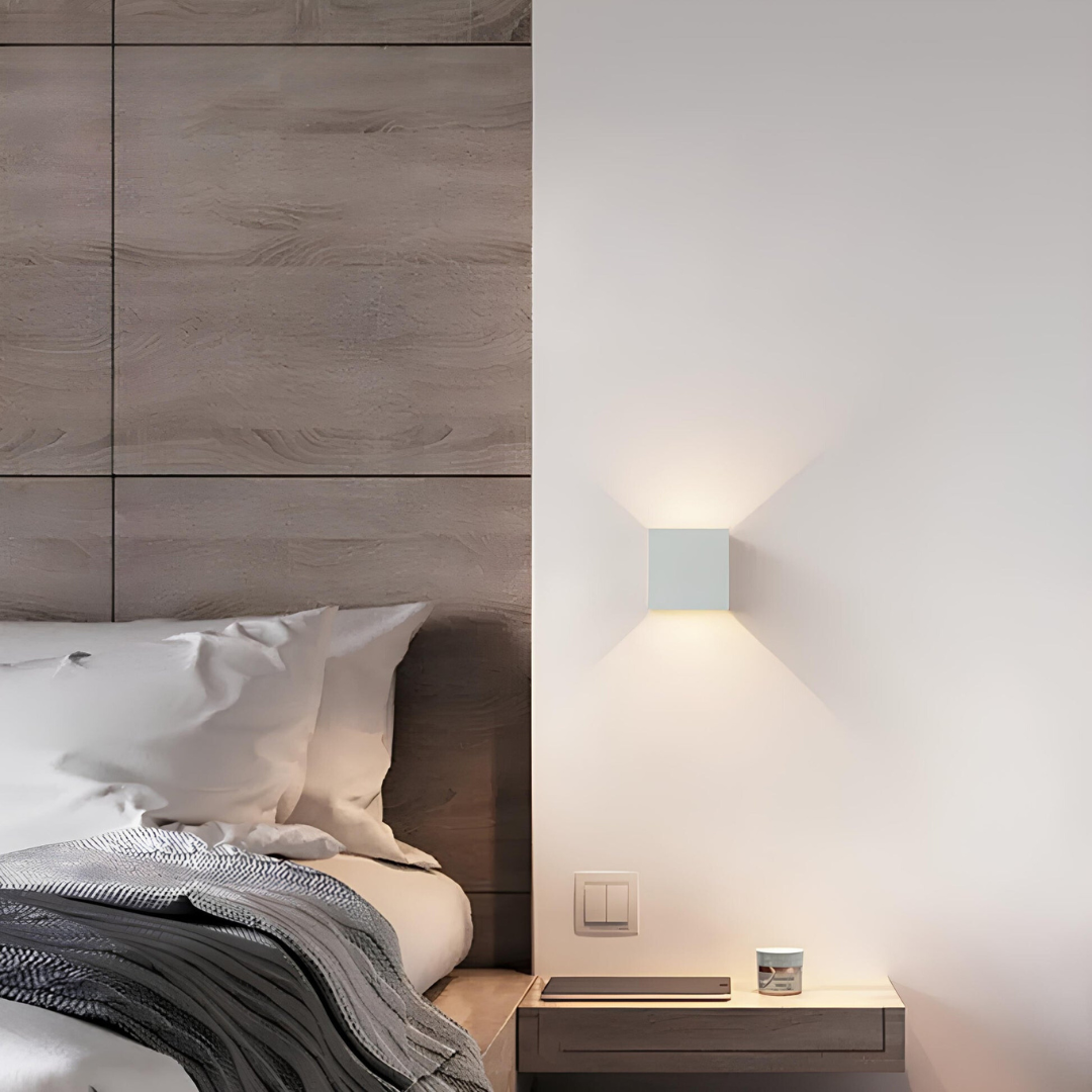 White cube wall lamp beside a bed with a wood-panelled headboard, above a floating bedside shelf holding a tablet and small jar. 6