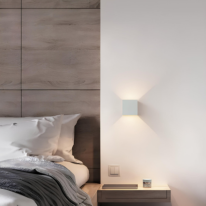 White cube wall lamp beside a bed with a wood-panelled headboard, above a floating bedside shelf holding a tablet and small jar. 6