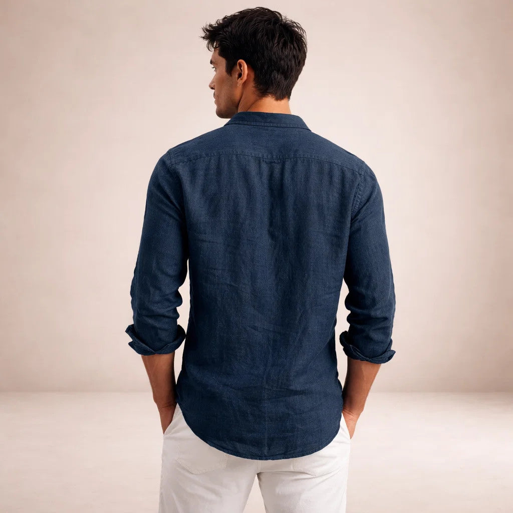 Back view of a model in a navy linen shirt with rolled sleeves and hands in the pockets of white trousers. 13