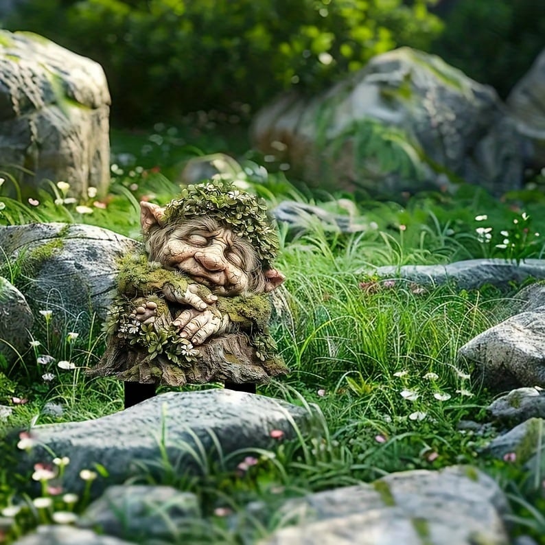 Small sleeping troll with a leafy crown and folded hands, staked in grass between mossy grey rocks. 16
