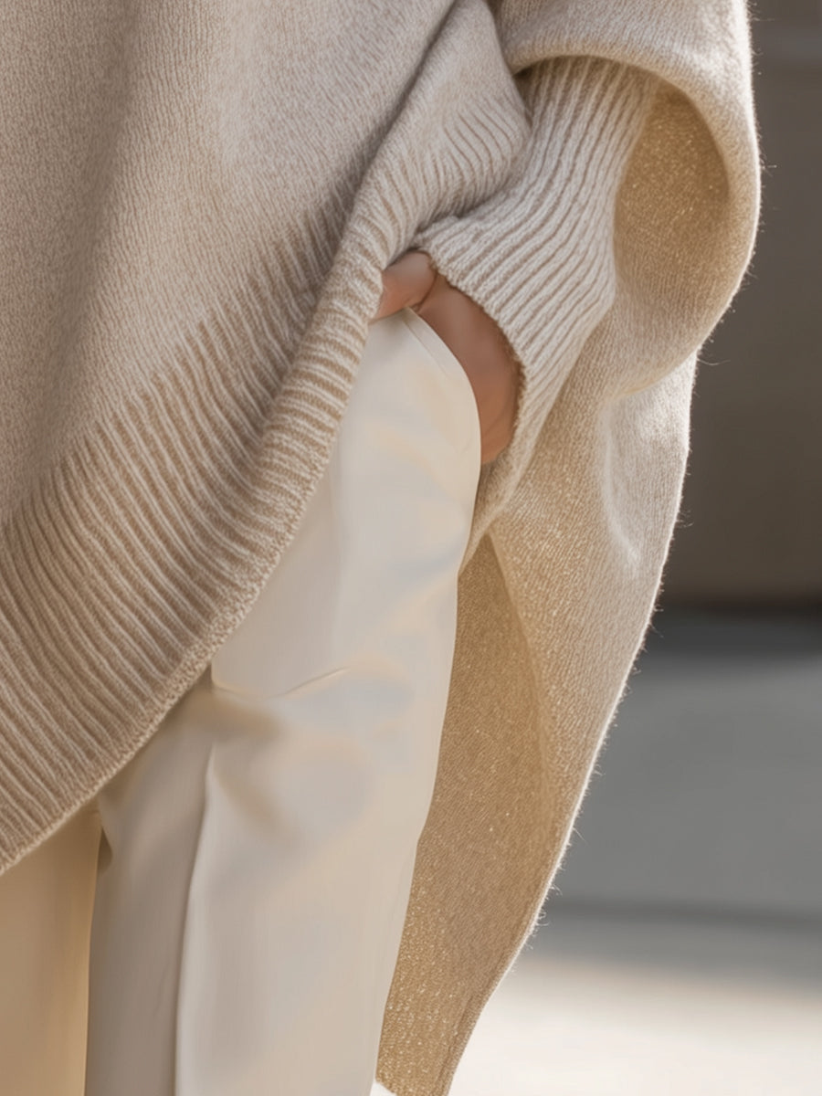 Close-up of the cream poncho's side slit, ribbed cuff and hem, with a hand in a trouser pocket 6