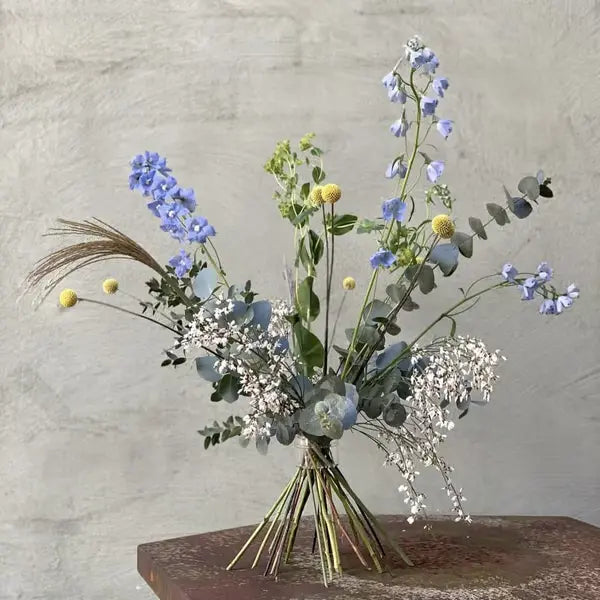 Upright hand-tied bouquet of blue delphiniums, yellow craspedia and eucalyptus with spiralled stems, standing on a rusty table against a grey wall. 12
