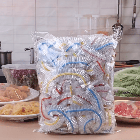 Large clear bag packed with clear stretchy covers with blue, yellow and red bands, standing on a kitchen counter beside covered dishes. 1