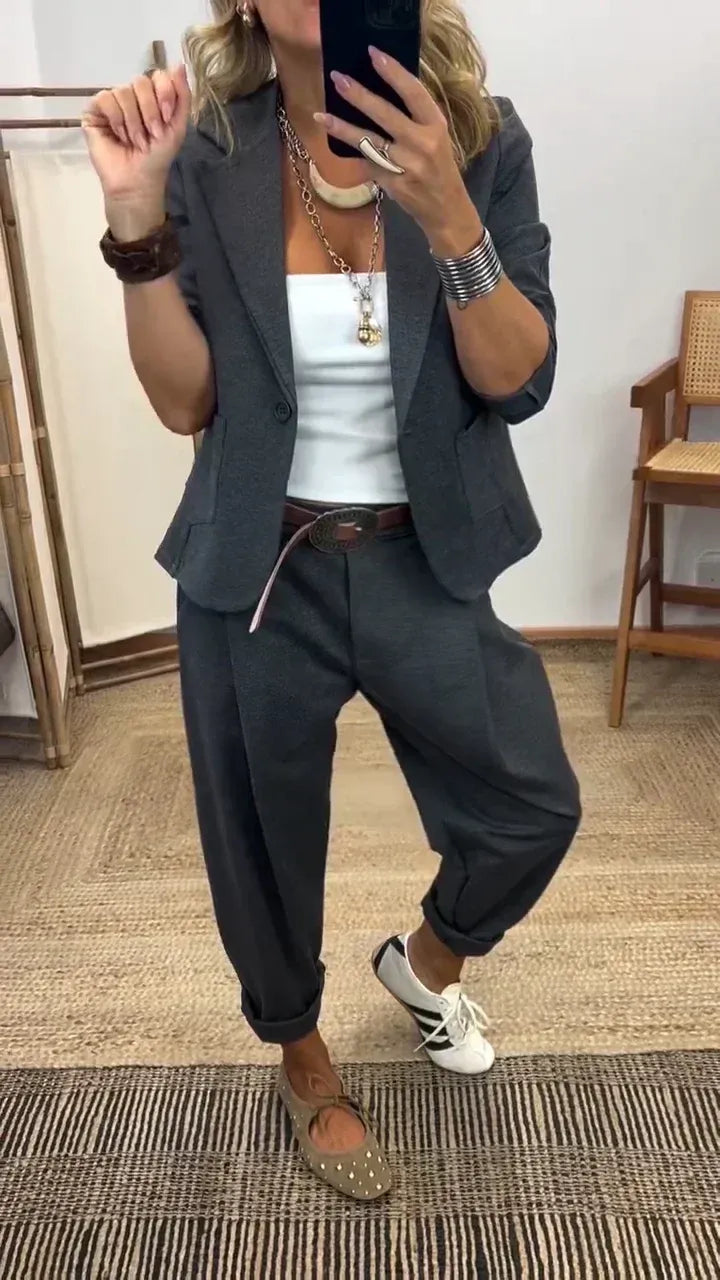 Woman in the dark grey blazer and rolled-cuff trousers over a white top, taking a mirror selfie with a brown belt. 1