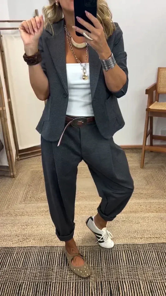 Woman in the dark grey blazer and rolled-cuff trousers over a white top, taking a mirror selfie with a brown belt. 1