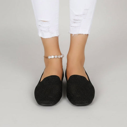 Front view of black slip-on shoes worn by a model in white ripped jeans and a pearl anklet. 2