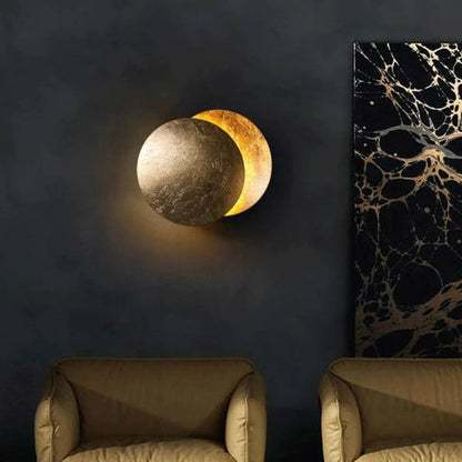 Gold moon wall lamp on a dark wall beside marble-style artwork, above two tan armchairs 3