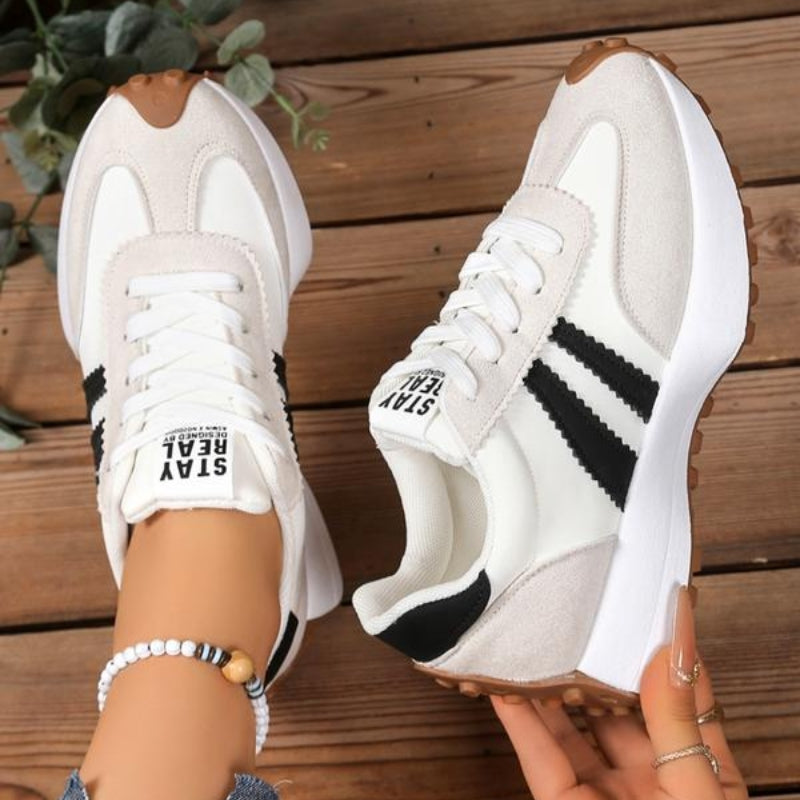 Cream and white trainers with black stripes, one worn and one held in hand above wooden boards 10