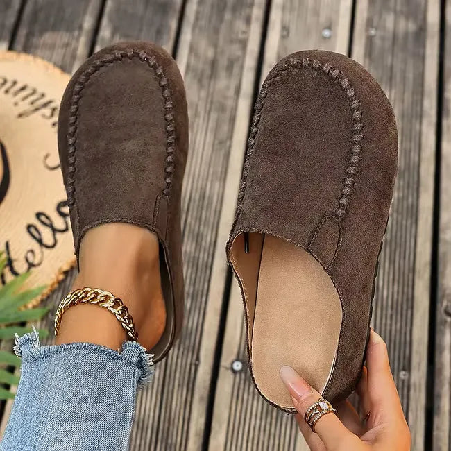Dark brown suede-look slip-ons seen from above, one on the foot and one held open to reveal the pale insole. 3