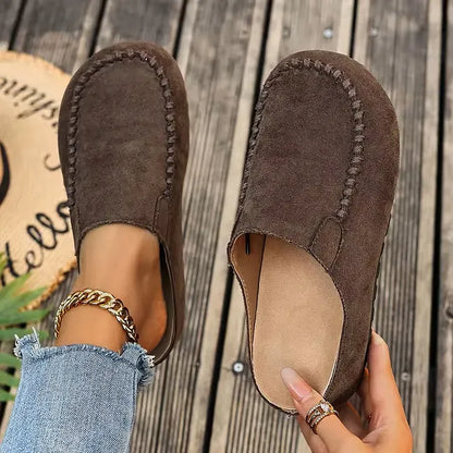 Dark brown suede-look slip-ons seen from above, one on the foot and one held open to reveal the pale insole. 3