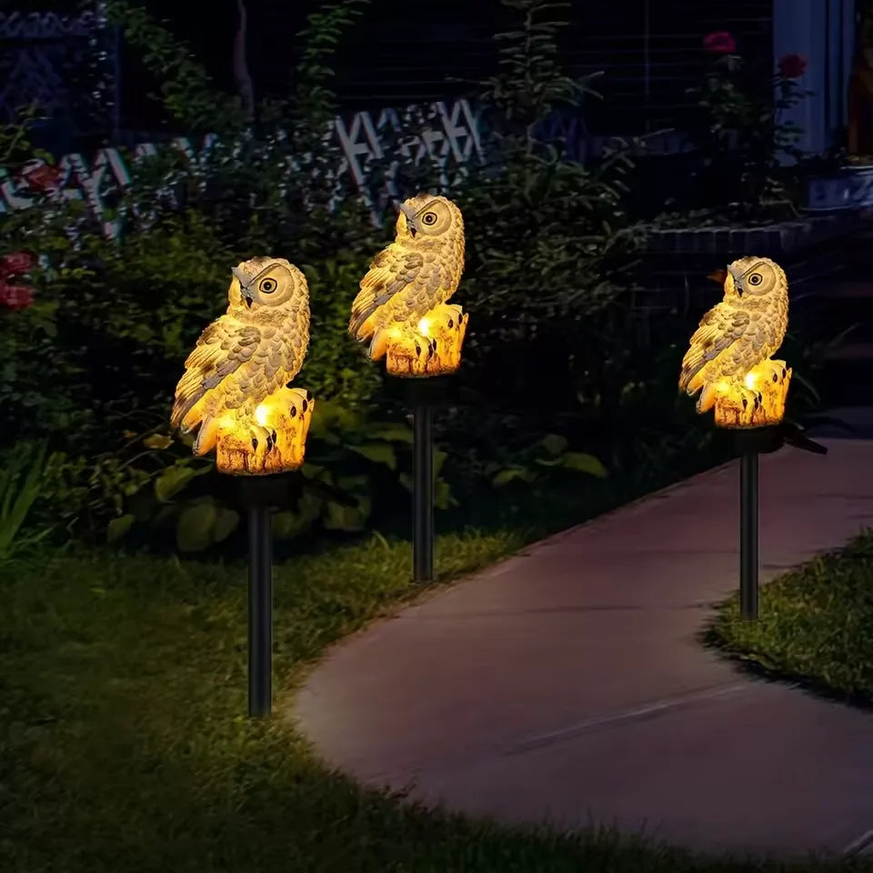 Three lit owl lights in a row on stakes beside a curving garden path at night. 9