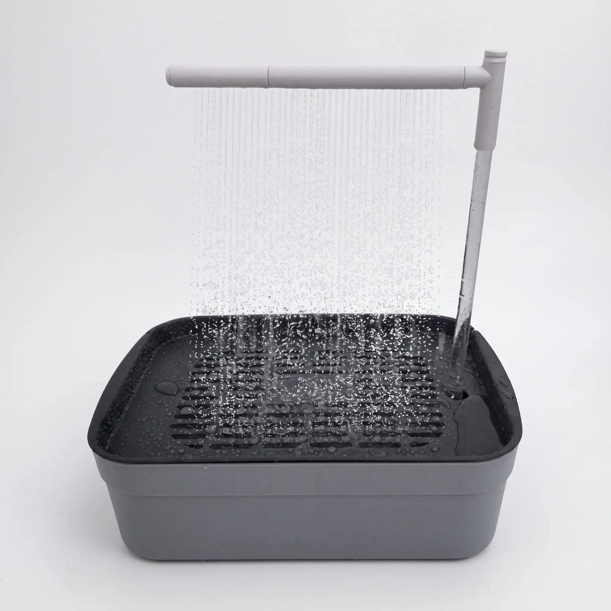 Grey bird bath with a dark slatted tray and a white overhead arm spraying a fine curtain of water, shown against plain white. 1