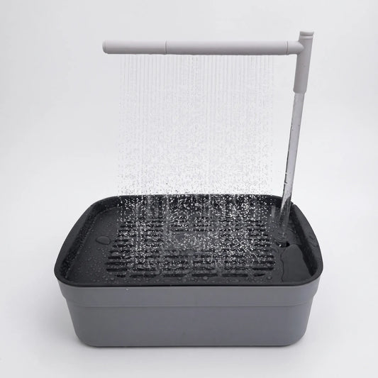 Grey bird bath with a dark slatted tray and a white overhead arm spraying a fine curtain of water, shown against plain white. 1