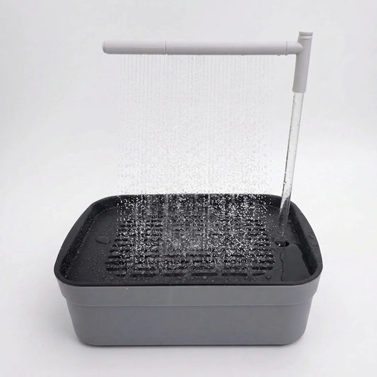 Grey rectangular bird bath on a plain white background, with an overhead arm spraying a curtain of water onto the slatted tray. 1
