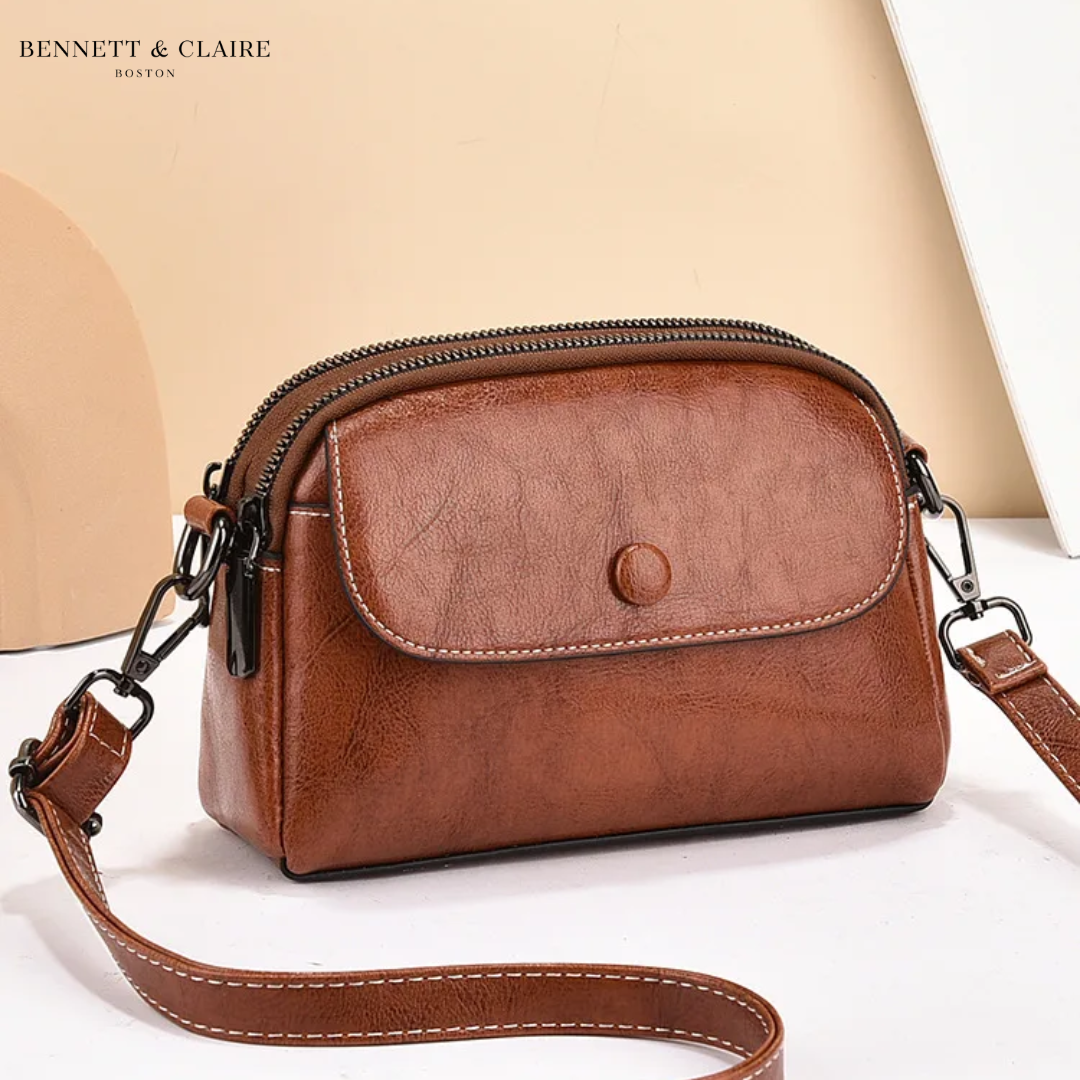 Tan-brown crossbody bag with a stitched front flap, press-stud fastening, twin zip top and dark metal clasps, shown on a pale studio set. 1