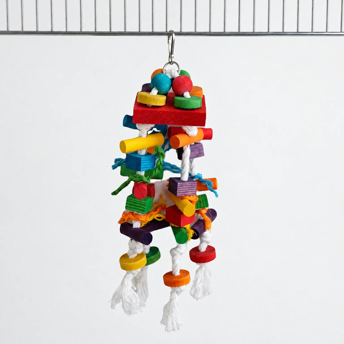 Toy of dyed wooden blocks, beads and knotted white cotton rope hanging from a metal clip on a cage bar against a plain background. 1