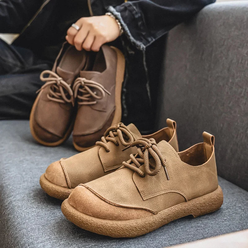 Tan lace-up shoes on a grey sofa while a person behind holds brown shoes 13
