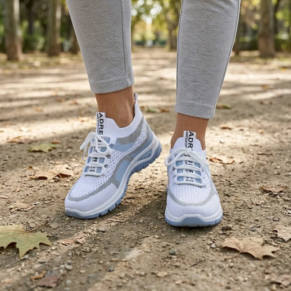 Pair of white and pale blue mesh trainers worn by a model in grey leggings, walking on a leafy park path. 2