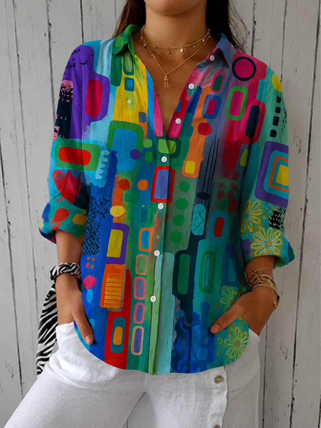 Front view of the blouse in bright rainbow shades of green, purple and red with a classic collar, worn with white trousers. 2
