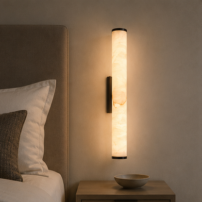 Illuminated marble wall light with dark end caps beside a bed headboard, above a wooden bedside table holding a shallow bowl. 9