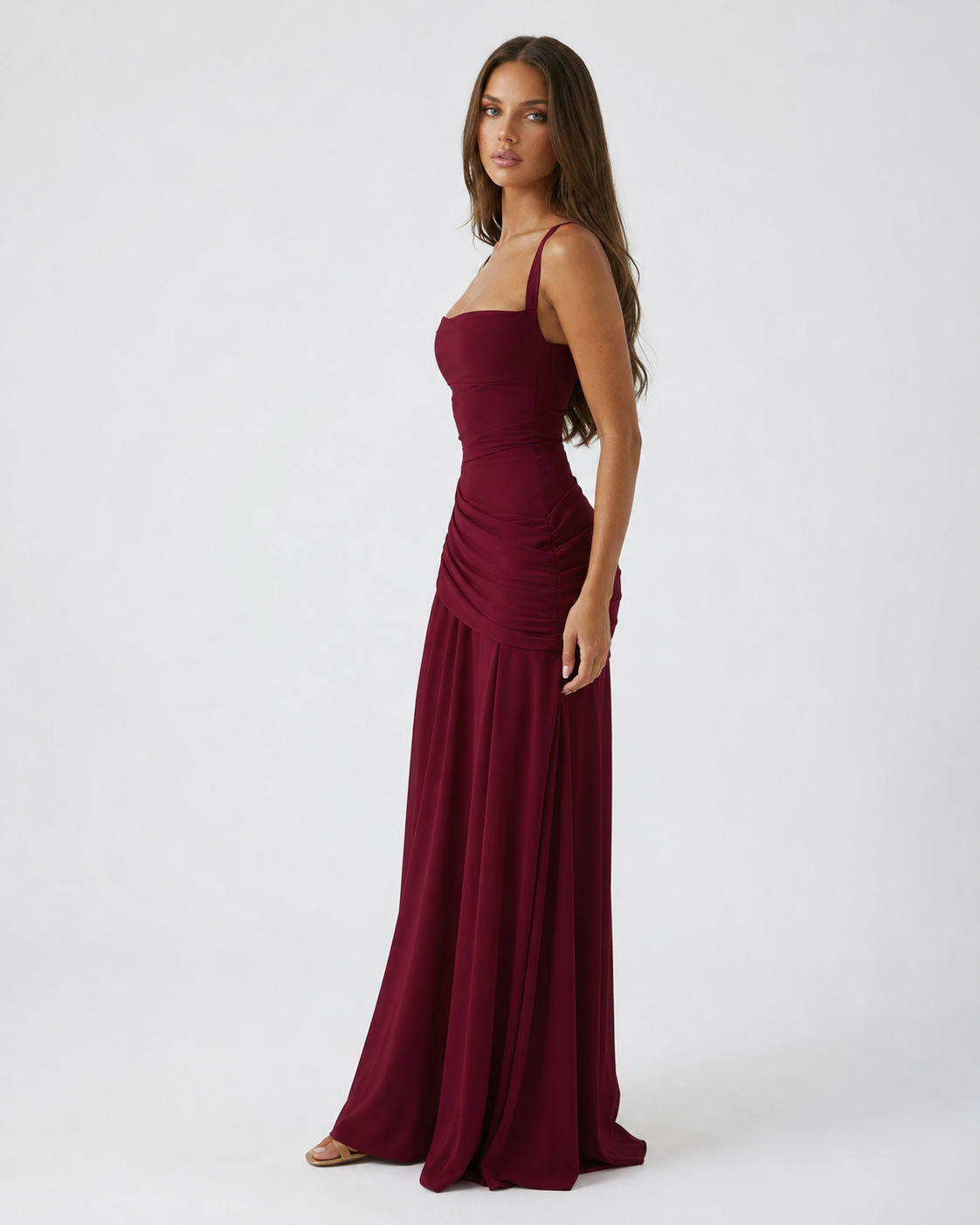 Burgundy dress seen from the side on a model, showing the thin strap, ruched waist and long skirt. 17