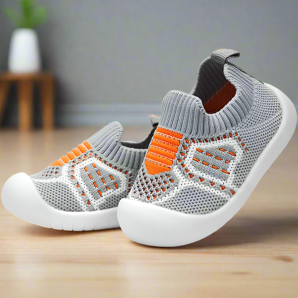 Grey knit baby shoes with orange stripes, mesh panels, heel pull tab and white rubber soles, angled on a wooden floor. 16