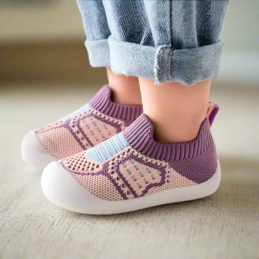 Side view of purple and pink knit baby shoes with ribbed cuffs and white rubber soles, worn on a carpeted floor. 15