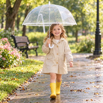 Girl in a beige raincoat and yellow wellies walks along a wet park path holding the clear umbrella. 3