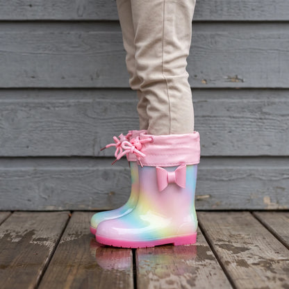 Child in beige trousers wearing rainbow wellies with a pink drawstring cuff, tied laces and bow, standing on a wet wooden deck. 3