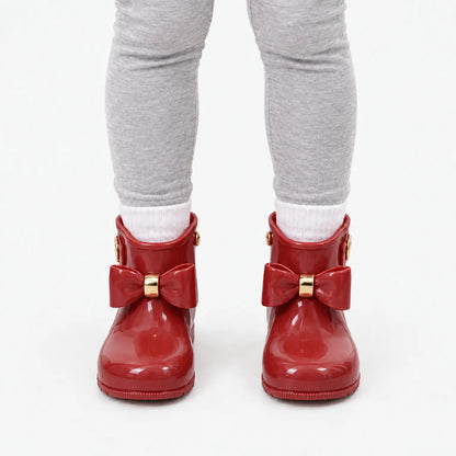 Front view of a child wearing glossy red bow wellies with white socks and grey leggings. 10