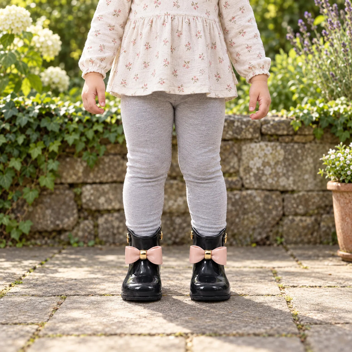Child standing in a sunny garden wearing black glossy wellies with pink bows, grey leggings and a floral top. 3