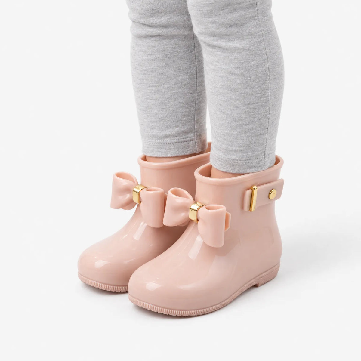 Angled view of a child's legs in pink glossy wellies with bows and a gold-buckled side strap. 5