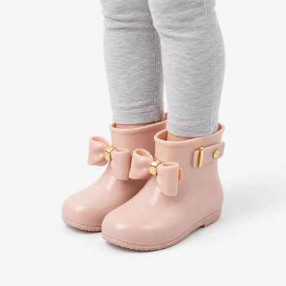 Angled view of a child's legs in pink glossy wellies with bows and a gold-buckled side strap. 5