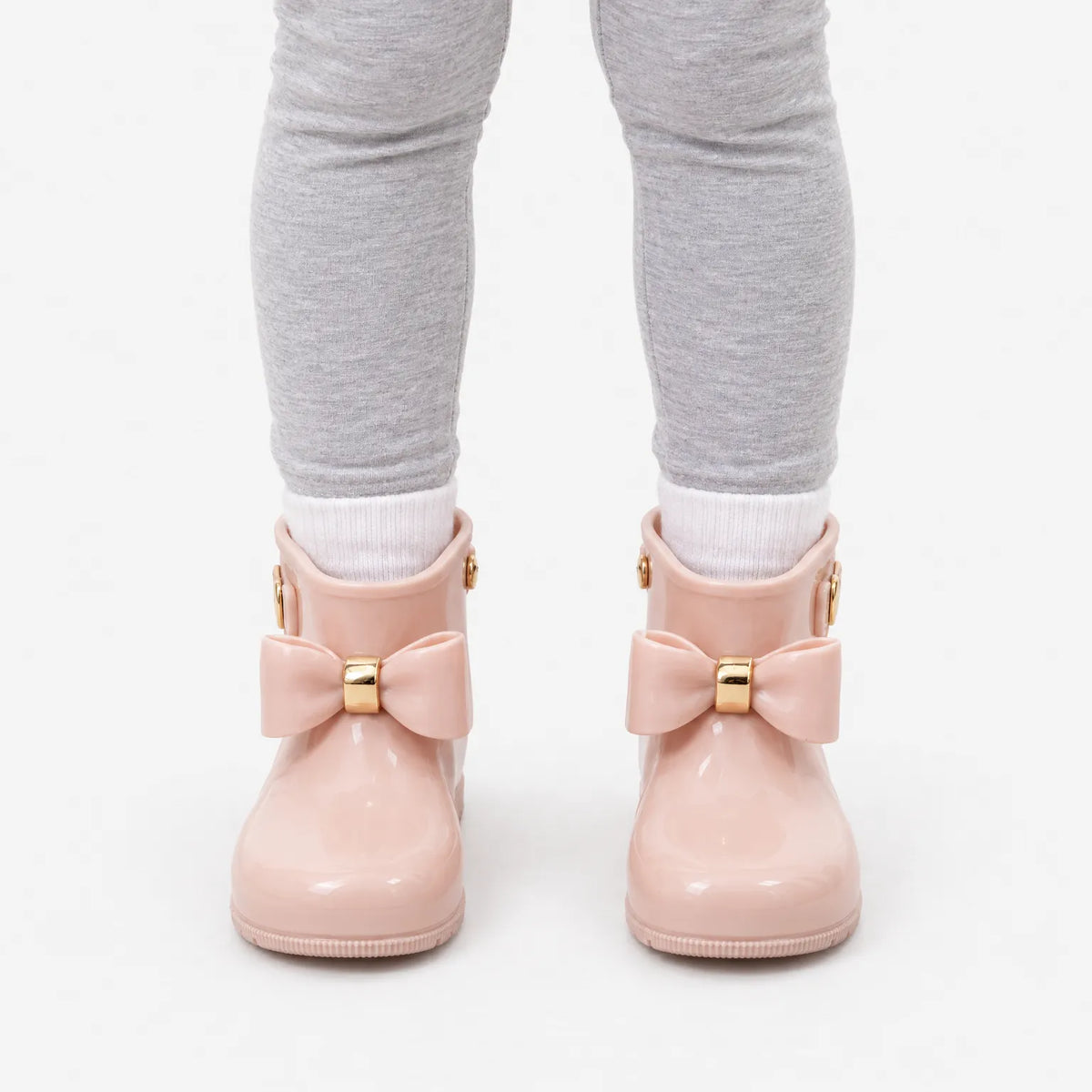 Front view of a child wearing pink bow wellies with white socks and grey leggings against a white background. 7