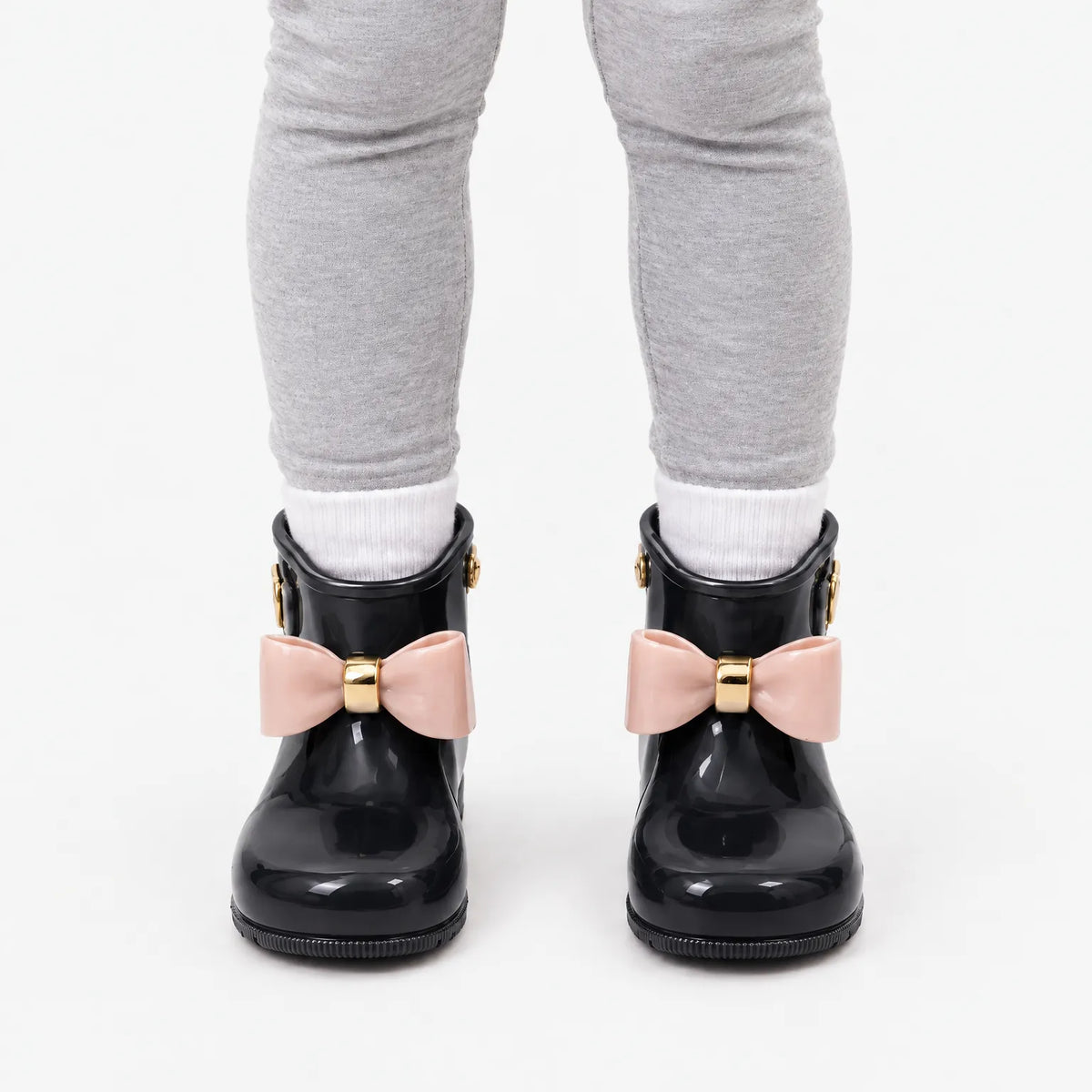 Front view of a child wearing black glossy wellies with pink bows, white socks and grey leggings. 9