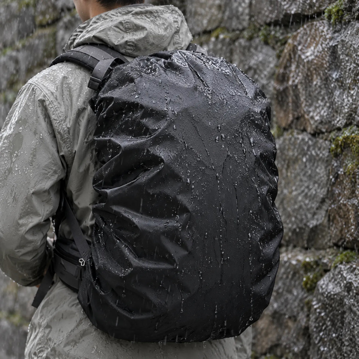Hiker in a grey waterproof jacket seen from behind in heavy rain, wearing a black rucksack cover beaded with water against a wet stone wall. 1