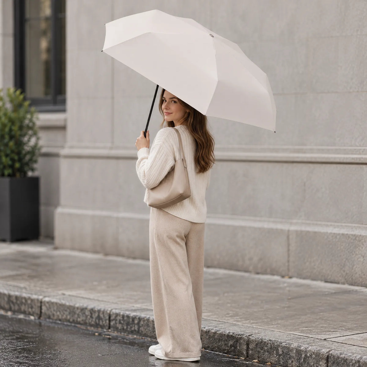 Woman in cream knitwear and beige trousers holds the open cream umbrella over her shoulder on a wet city pavement. 1
