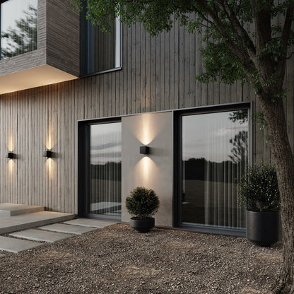 Black cube wall lamps lighting up and down on a timber-clad house exterior at dusk, near large windows and potted plants. 3