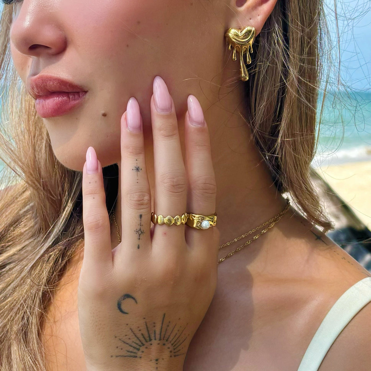 Close-up of a woman wearing a gold melting heart earring with gold rings and delicate necklaces, standing on a beach. 4