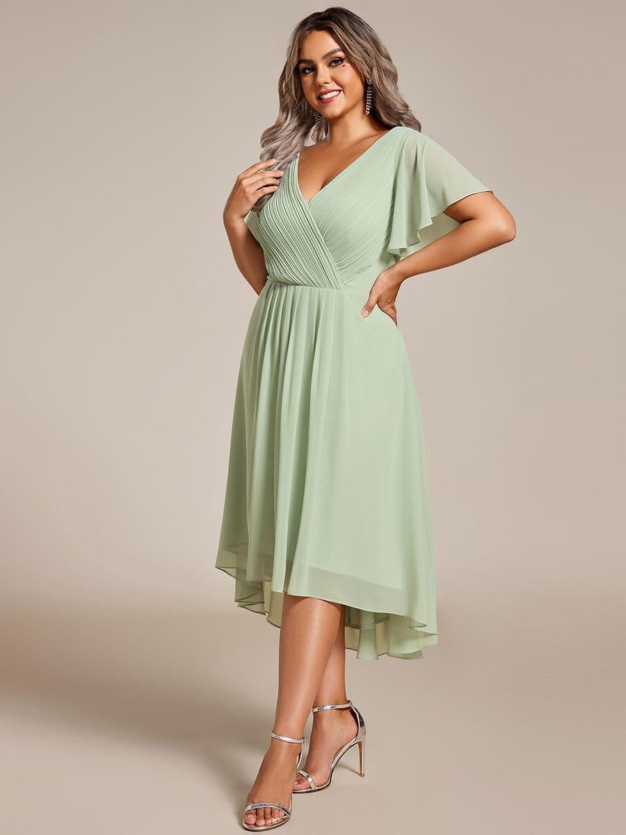 Plus-size model in a sage green chiffon dress with pleated wrap bodice and flutter sleeves, posed three-quarter view with silver heels. 81