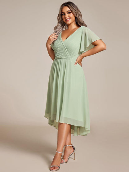 Plus-size model in a sage green chiffon dress with pleated wrap bodice and flutter sleeves, posed three-quarter view with silver heels. 81