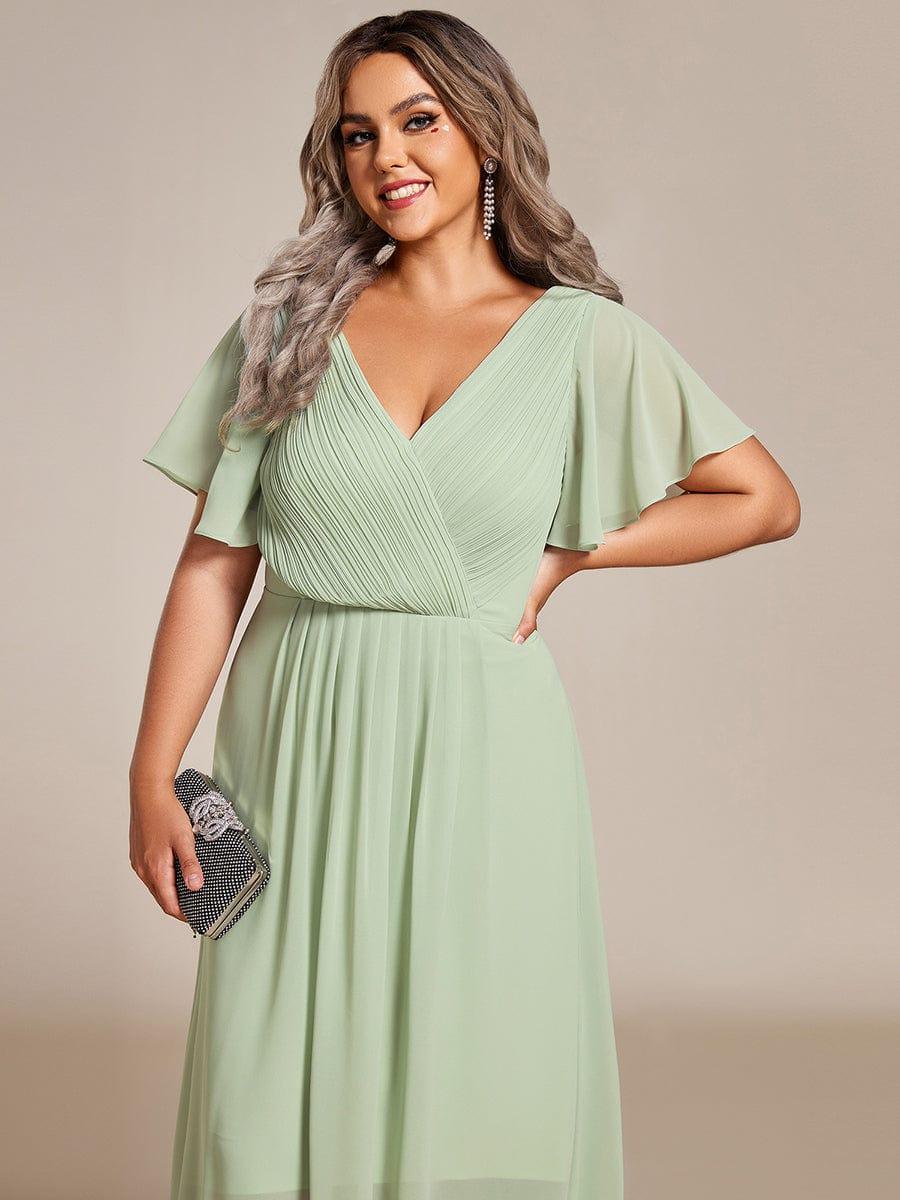 Close-up of the sage green pleated wrap V-neck bodice and flutter sleeves on a plus-size model holding a sparkly clutch. 83