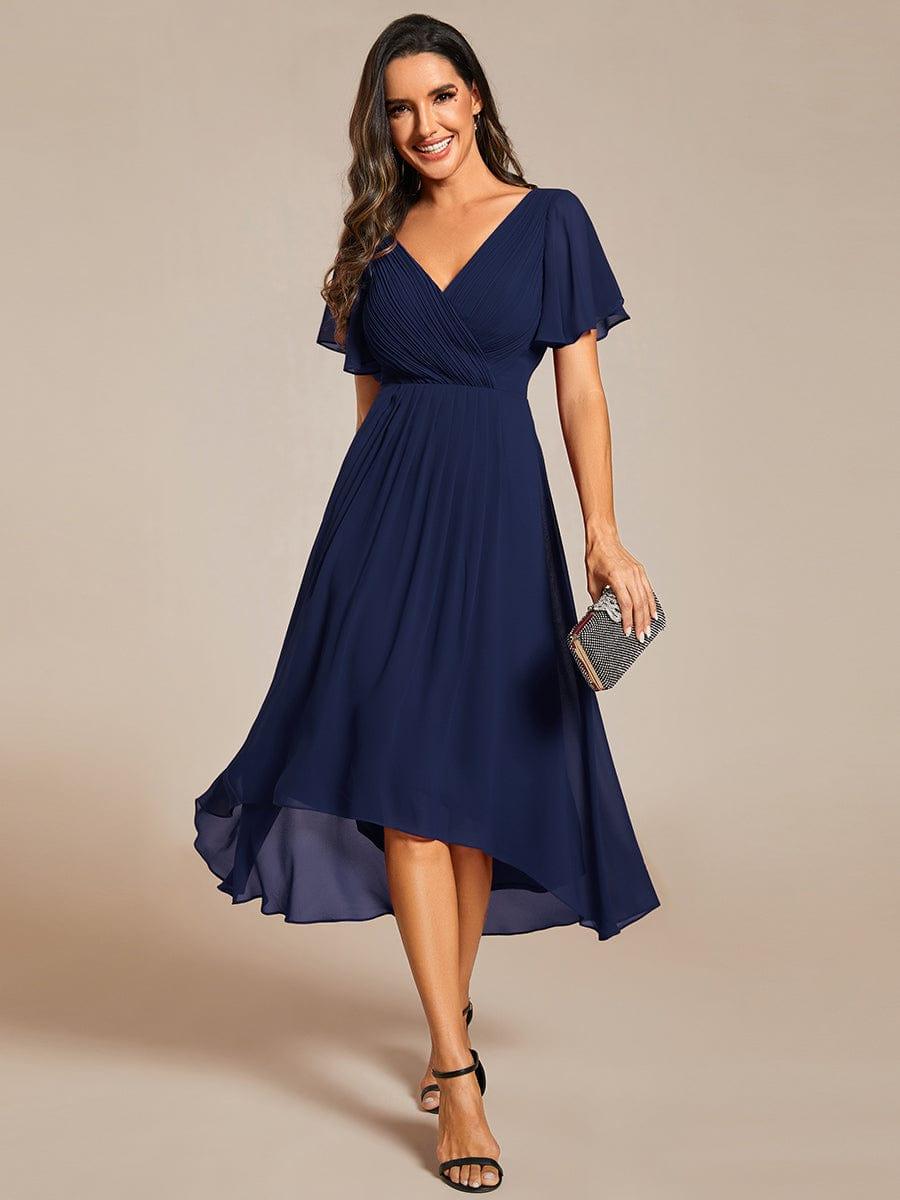 Model wearing the navy chiffon high-low dress from the front, smiling, with a glittery clutch and black strappy heels. 1