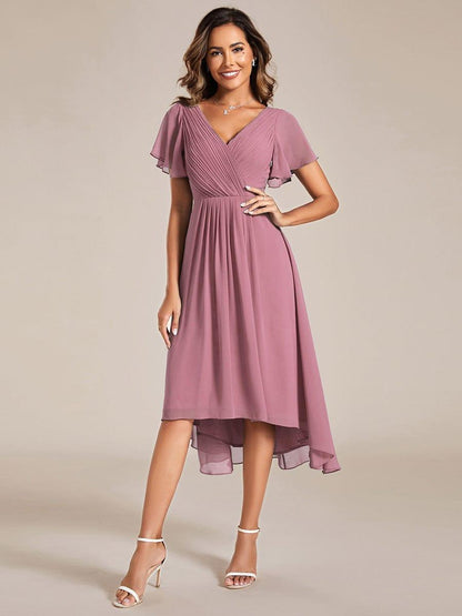 Model in the mauve-pink high-low chiffon dress with pleated V-neck and flutter sleeves, wearing white heels, hand on hip. 58