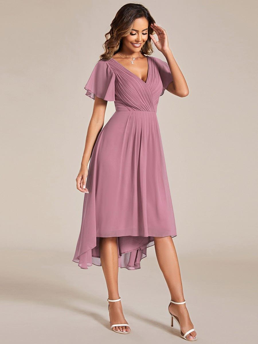 Dusky mauve pink chiffon dress with pleated V-neck and high-low hem, worn with white heeled sandals, model touching her hair. 62