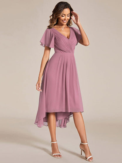 Dusky mauve pink chiffon dress with pleated V-neck and high-low hem, worn with white heeled sandals, model touching her hair. 62